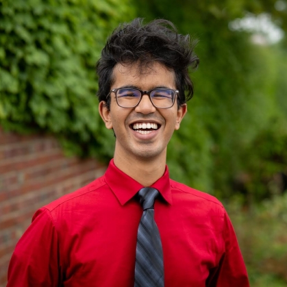 Young man with black hair and glasses smiles broadly wearing a red shirt with a black and gray stripped tie. He is standing in front of a brick wall and bushes.