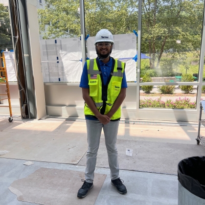 Male college student is on a construction site wearing a hard hat, yellow vest, and light colored pants.