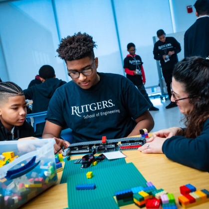 Student with black hair and glasses wearing a black Rutgers T-shirt works at a table with young students on a Lego project.