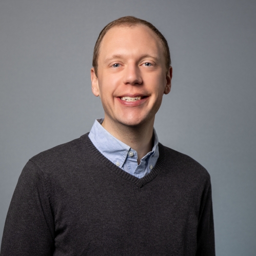 head shot of male wearing a brown pullover sweater over a blue button down shirt.