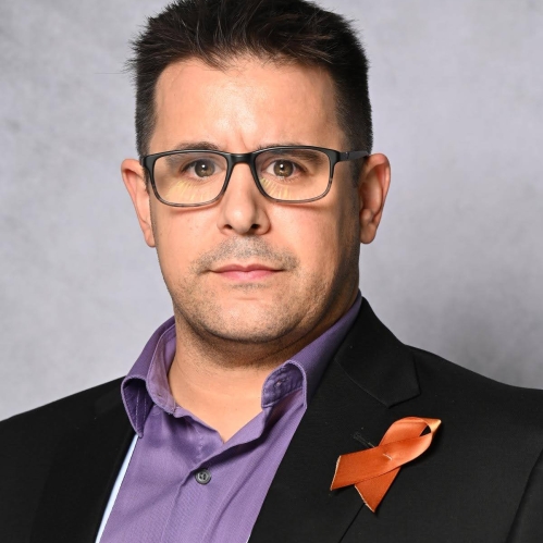 Headshot of male with short dark hair with eyeglasses, wearing a dark blue suit jacket with a purple button down shirt and an orange cancer ribbon pinned on his lapel.