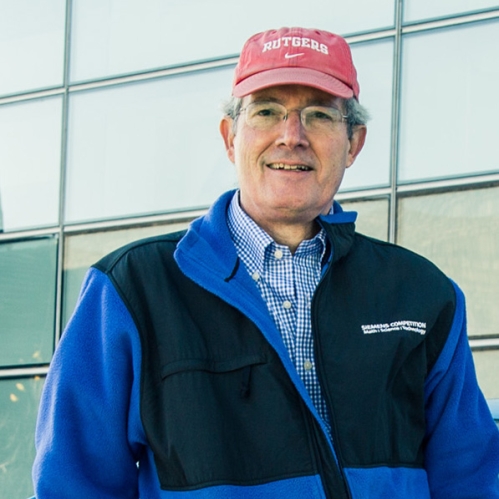 Male leaning on car, wearing eyeglasses, a red baseball cap, blue and black zip up jacket.