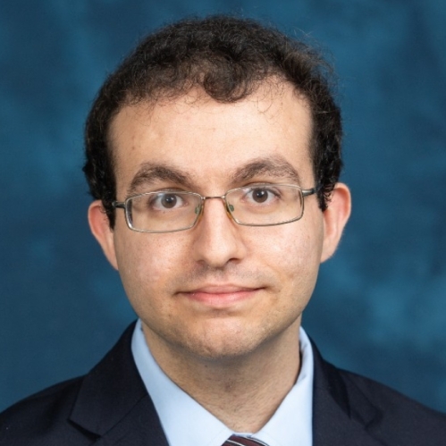 Head shot of male with dark brown, short hair wearing eyeglasses, a dark suit with a light blue button down shirt with a patterned tie. and
