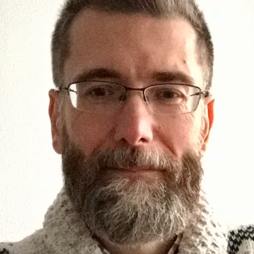 Headshot of male with short light brown and grey hair with facial hair and wearing eyeglasses and a beige sweater.