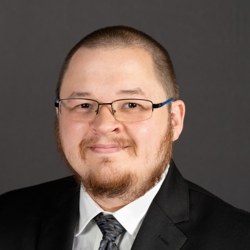 Headshot of male with a buzz haircut, facial hair, wearing a black suit, with a white shirt, and a dark tie.
