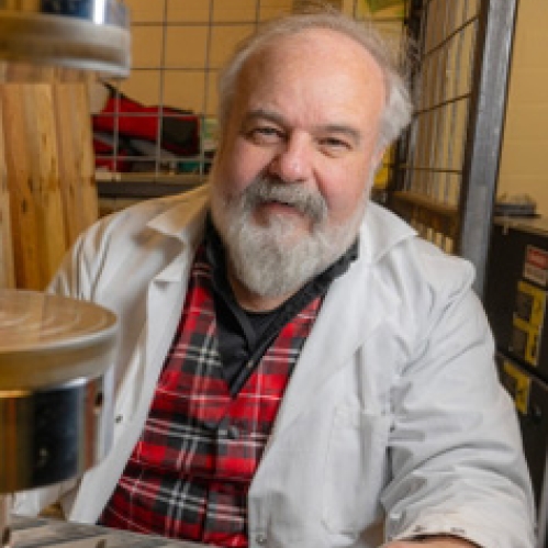 Balding male with grey beard and moustache sitting in lab wearing a white lab coat over a red and black plaid shirt.