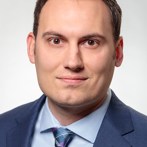 Headshot of male with short brown hair, wearing a blue suit, light blue shirt, and a patterned blue tie.