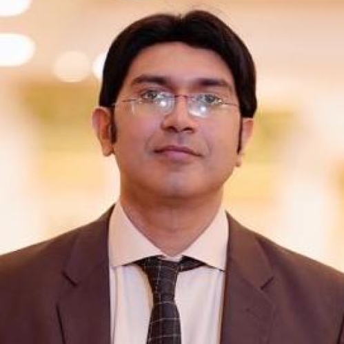 Headshot of male with black hair, wearing eyeglasses and brown suit jacket with a tan colored shirt and brown tie.
