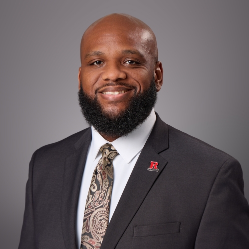 Headshot of balding black male with black beard and moustache, wearing a black suit, white button down shirt, patterned tie, and a R lapel pin.