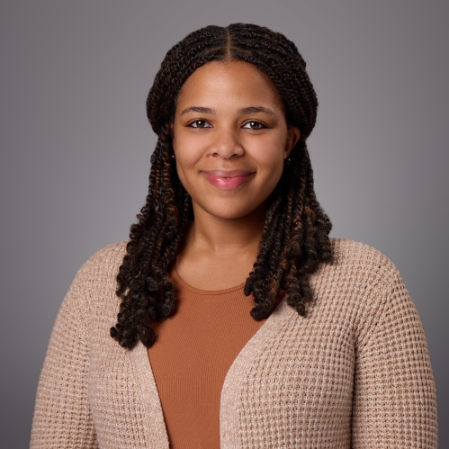 Headshot of black woman with long black hair wearing a brown shirt under a tan sweater. and brown shirt