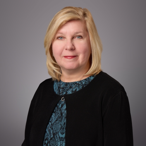 Headshot of female with shoulder length blonde hair, wearing a green patterned blouse under a black sweater.