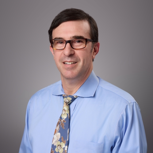 Headshot of male with short brownhair and eyeglasses, wearing and light blue shirt and patterned blue tie.