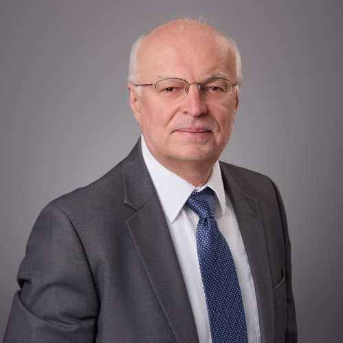 Headshot of male with white hair and eyeglasses wearing a grey suit, white shirt, and blue patterned tie.