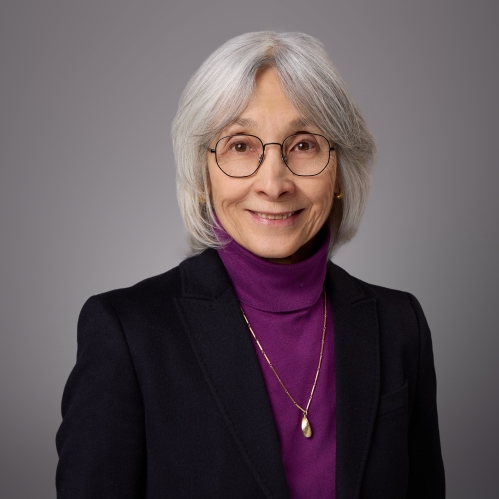 Headshot of female with short grey hair wearing a purple turtleneck, black jacket and a necklace.