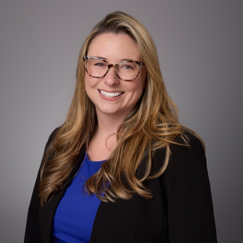 Headshot of woman with long lilght brown hair and eyeglasses wearing a blue blouse under a black sweater. .