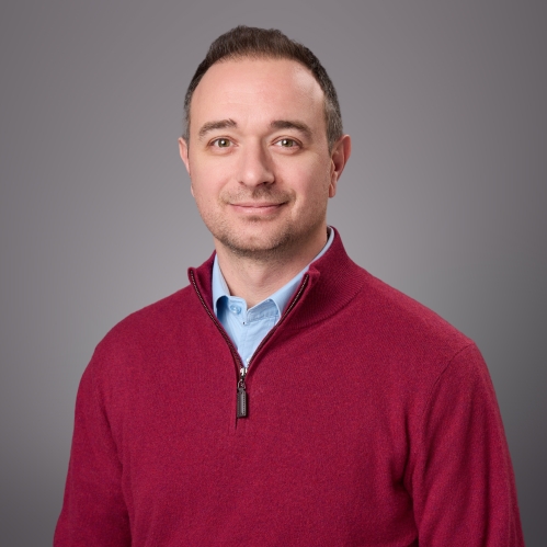 Headshot of male wearing a light blue shirt under a burguny pullover. light blue shirt