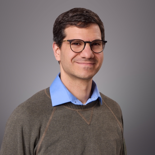 Headshot of male with eyeglasses wearing a blue collared shirt underneath a brown pullover shirt.