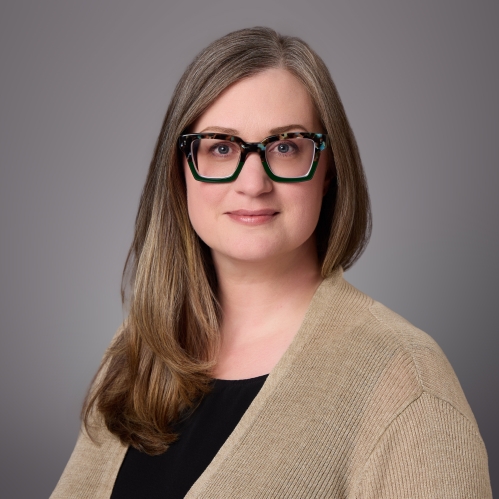 Woman with long brown hair with eyeglasses wearing a blouse under a light brown sweater.