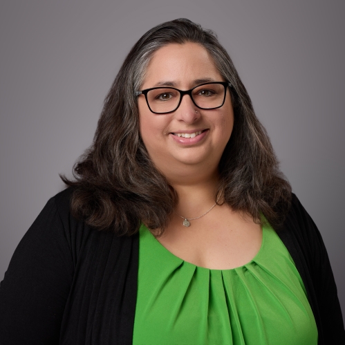 Headshot of woman with long brown hair and eyeglasses wearing a green blouse underneath a black sweater.