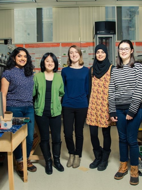 Group of five young women in an engineering lab all standing in a line looking at the camera, smiling.