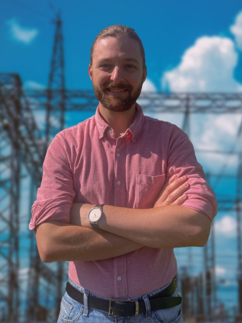 head shot of male standing in front of power grid with arms folded, with short blonde hair and moustache and beard, wearing a salmon color button down shirt