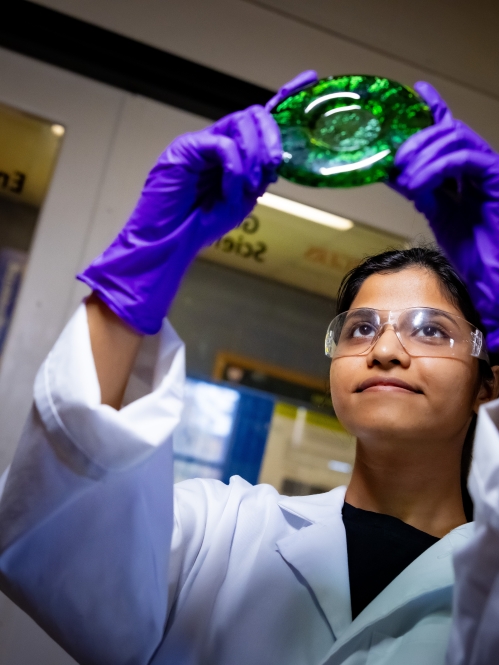 Woman wearing a lab coat and purple safety gloves holds up a round piece of green glass and looks at it.