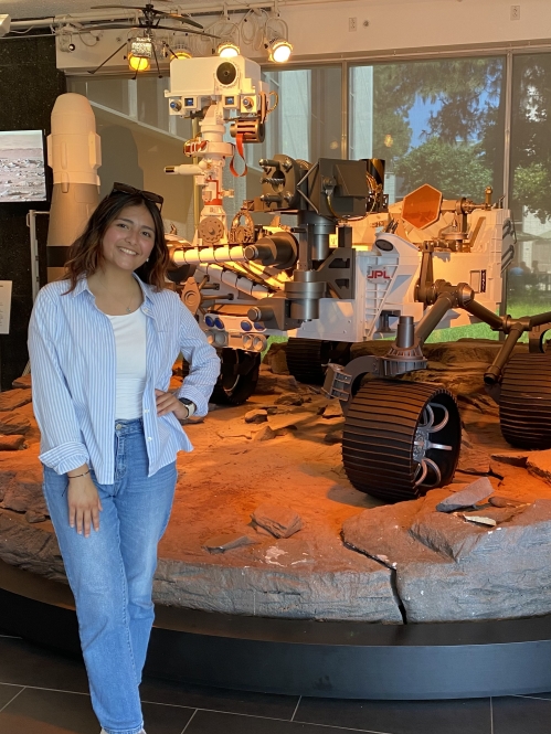 Female young woman poses with her hand on her hip, wearing jeans and a light blue shirt. She has long dark hair and she is standing in front of a lunar rover.