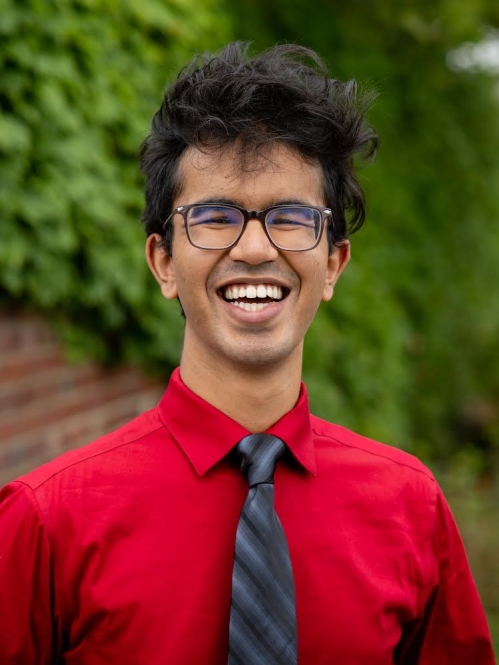 Young man with black hair and glasses smiles broadly wearing a red shirt with a black and gray stripped tie. He is standing in front of a brick wall and bushes.