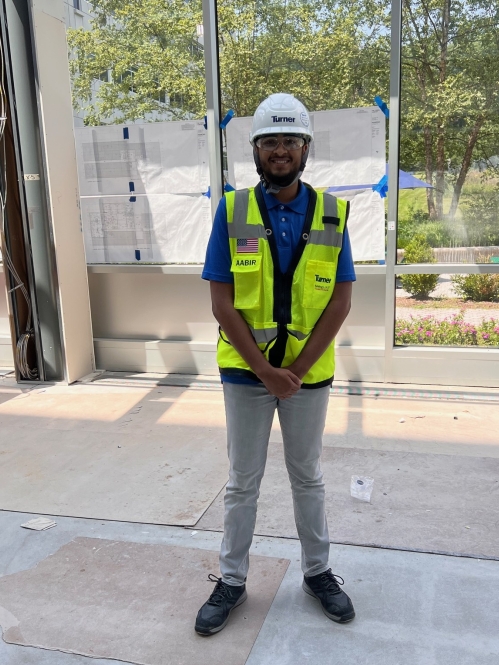 Male college student is on a construction site wearing a hard hat, yellow vest, and light colored pants.