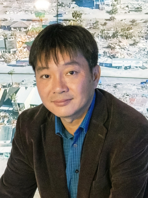 Head shot of Asian male with short black hair wearing a dark brown suit jacket with a blue button down shirt. 