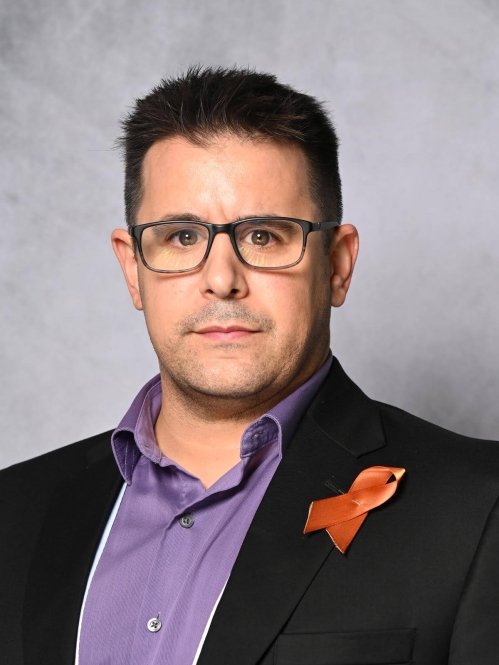 Headshot of male with short dark hair with eyeglasses, wearing a dark blue suit jacket with a purple button down shirt and an orange cancer ribbon pinned on his lapel.