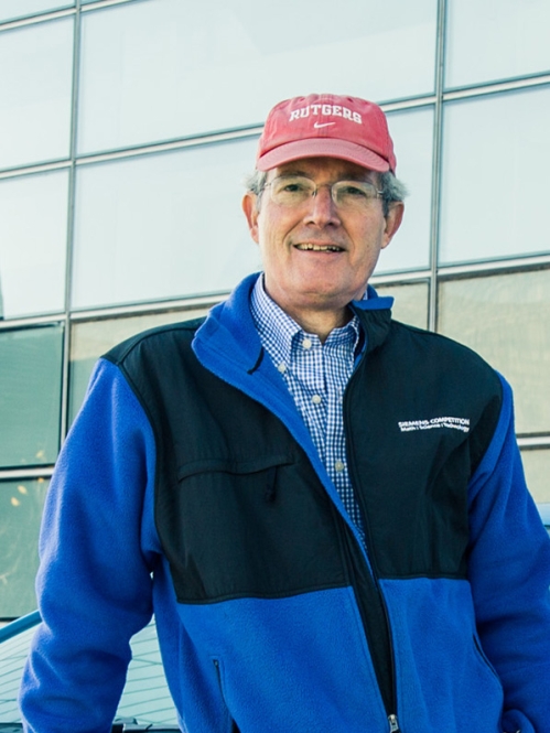 Male leaning on car, wearing eyeglasses, a red baseball cap, blue and black zip up jacket.