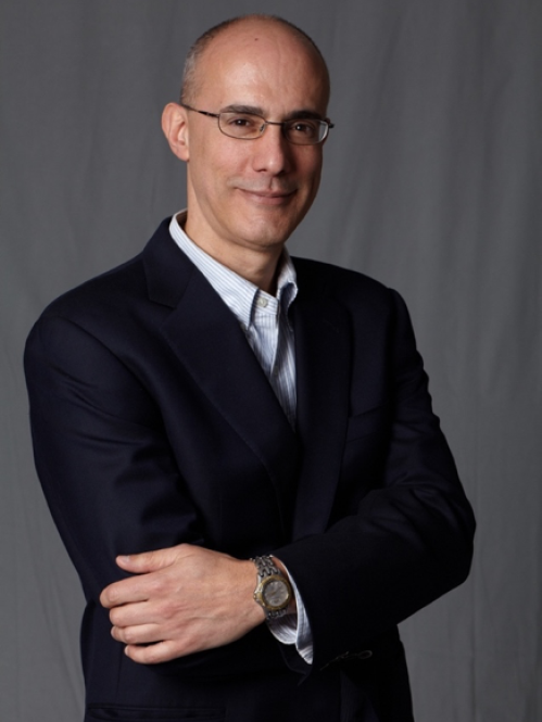Headshot of bald male with his arms crossed, wearing eyeglasses, a black suit and a button down striped blue and whtie shirt.