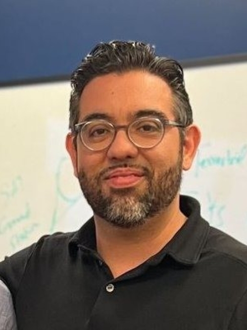 Headshot of male with short hair and facial hair, wearing eyeglasses and a black polo shirt.