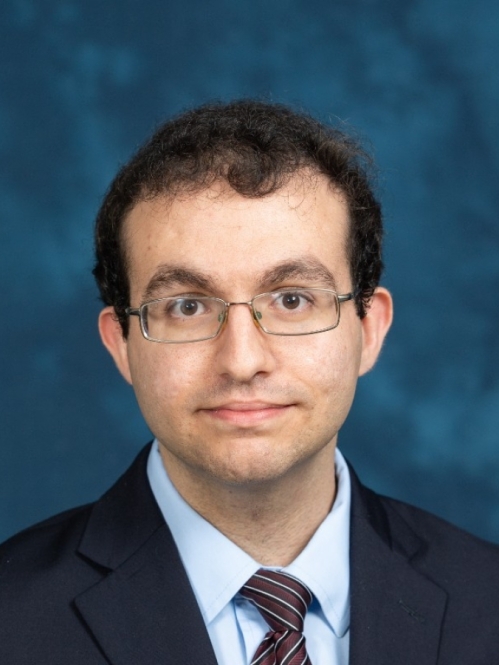 Head shot of male with dark brown, short hair wearing eyeglasses, a dark suit with a light blue button down shirt with a patterned tie. and