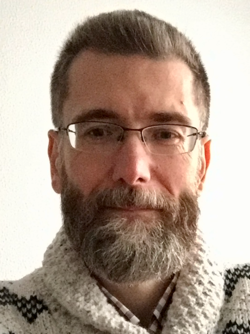 Headshot of male with short light brown and grey hair with facial hair and wearing eyeglasses and a beige sweater.