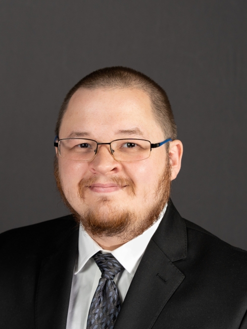 Headshot of male with a buzz haircut, facial hair, wearing a black suit, with a white shirt, and a dark tie.