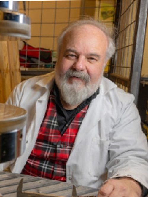  Balding male with grey beard and moustache sitting in lab wearing a white lab coat over a red and black plaid shirt.