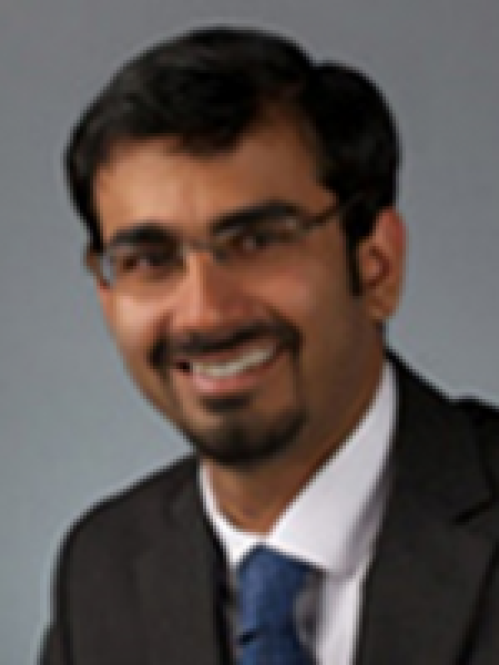 Headshot of male with short black hair and facial hair, wearing eyeglasses, a brown jacket, a white shirt, and a blue tie.t