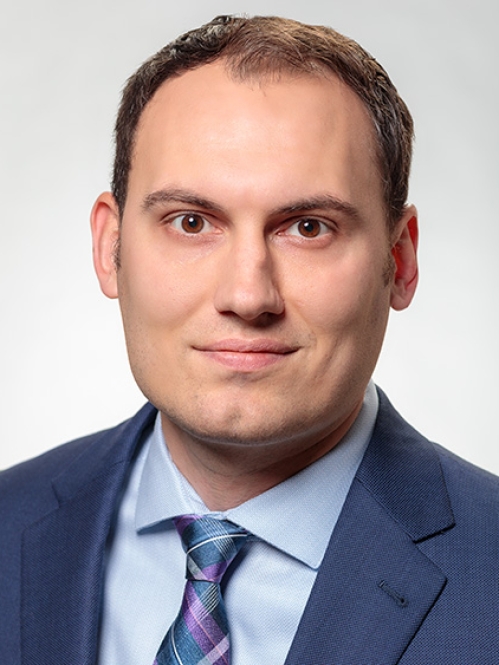 Headshot of male with short brown hair, wearing a blue suit, light blue shirt, and a patterned blue tie.