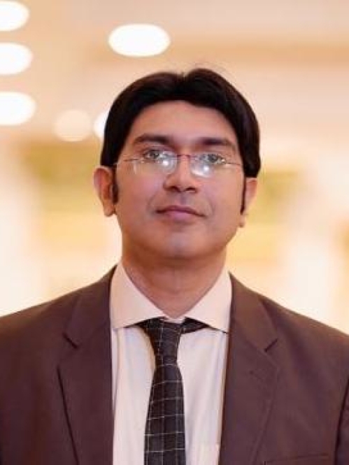 Headshot of male with black hair, wearing eyeglasses and brown suit jacket with a tan colored shirt and brown tie.