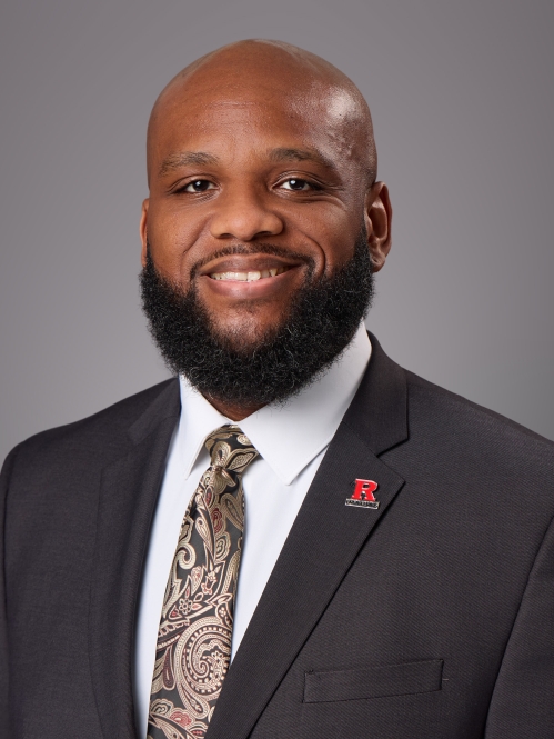 Headshot of balding black male with black beard and moustache, wearing a black suit, white button down shirt, patterned tie, and a R lapel pin. 
