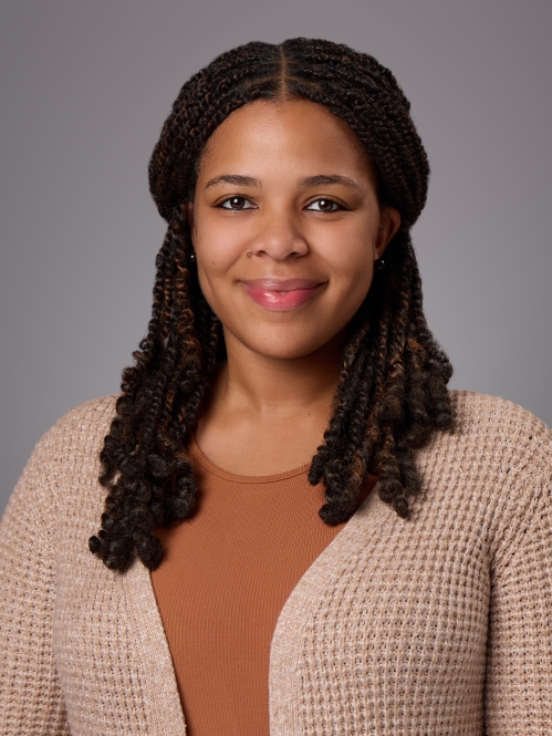 Headshot of black woman with long black hair wearing a brown shirt under a tan sweater. and brown shirt 