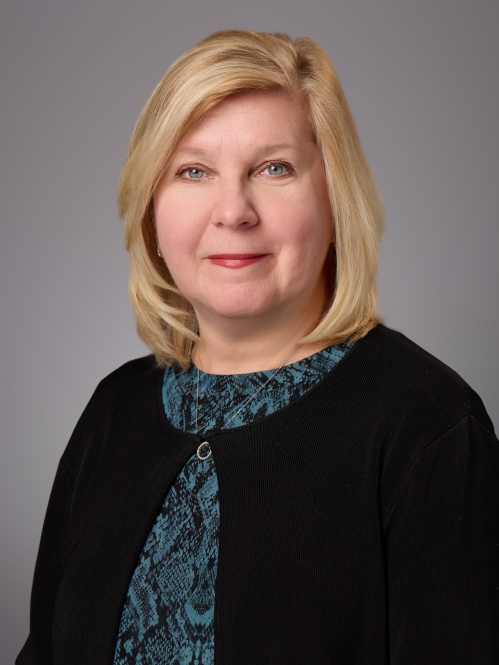 Headshot of female with shoulder length blonde hair, wearing a green patterned blouse under a black sweater.
