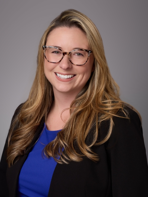 Headshot of woman with long lilght brown hair and eyeglasses wearing a blue blouse under a black sweater. .