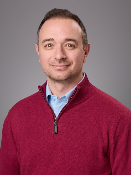 Headshot of male wearing a light blue shirt under a burguny pullover. light blue shirt