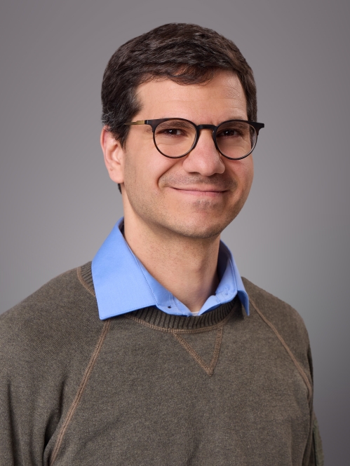 Headshot of male with eyeglasses wearing a blue collared shirt underneath a brown pullover shirt.