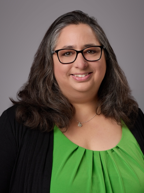 Headshot of woman with long brown hair and eyeglasses wearing a green blouse underneath a black sweater.