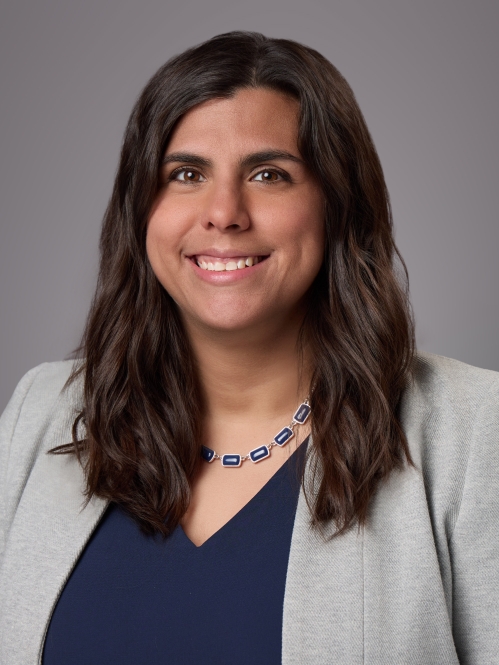 Headshot of female with long dark brown hair wearing a blue blouse under a grey jacket and a necklace