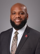 Headshot of balding black male with black beard and moustache, wearing a black suit, white button down shirt, patterned tie, and a R lapel pin.
